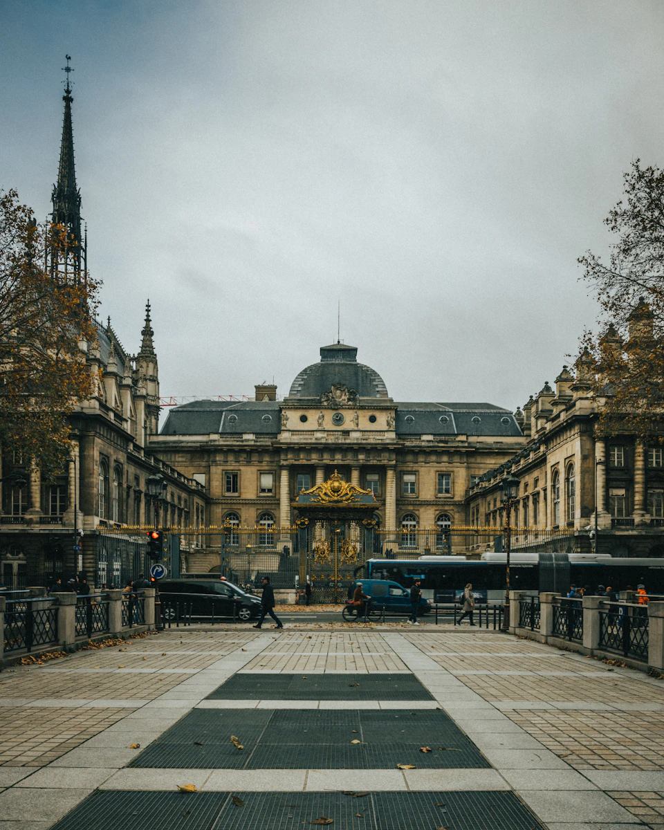 The image depicts a large, ornate building with a dome and multiple spires on its roof, situated within a city square. The building is surrounded by other structures, creating an urban landscape. A few people can be seen walking around the area, adding life to the scene. In front of the building, there are two traffic lights controlling the flow of vehicles in the vicinity.