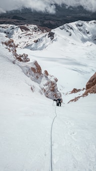 A lone climber ascends a steep, snow-covered mountain slope, using ice axes for support. The surrounding landscape is dominated by rugged, white peaks with patches of exposed rock. Clouds hang low in the sky, partially obscuring the horizon.