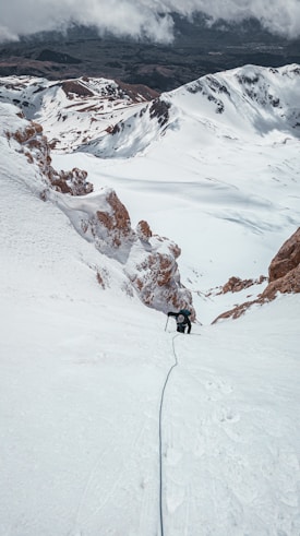 A lone climber ascends a steep, snow-covered mountain slope, using ice axes for support. The surrounding landscape is dominated by rugged, white peaks with patches of exposed rock. Clouds hang low in the sky, partially obscuring the horizon.