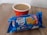 A cup of coffee in a red and white paper cup is placed on a wooden surface. In front of the cup, there is a blue packet of britannia Good Day butter cookies.