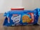 A packet of Britannia Good Day butter cookies is placed in front of a white and red cup. The packaging is predominantly blue with images of the cookies and the branding prominently displayed.