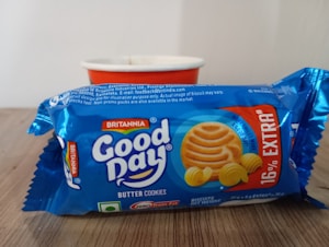 A packet of Britannia Good Day butter cookies is placed in front of a white and red cup. The packaging is predominantly blue with images of the cookies and the branding prominently displayed.