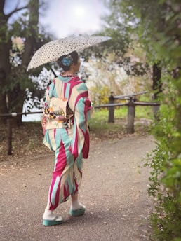 A person dressed in a vibrant kimono walks down a path surrounded by greenery, holding a patterned parasol. The kimono features bold stripes in pink, teal, and cream, complemented by traditional footwear. The scene appears serene and is set in a natural environment with trees and bushes lining the pathway.