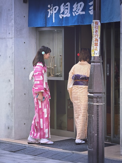 Two women wearing traditional kimonos stand in front of a shop entrance. One kimono is pink with white patterns, while the other is a light beige plaid. A blue cloth with Japanese characters hangs above the doorway. A vertical sign with colorful text is attached to the nearby post.