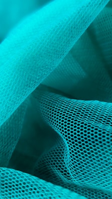 Waves of turquoise mesh fabric create an intricate and layered texture. The mesh features a repetitive hexagonal pattern, giving a sense of depth and dimensionality. The fabric is arranged in soft folds and curves, allowing light to play gently across its surface.