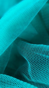 Waves of turquoise mesh fabric create an intricate and layered texture. The mesh features a repetitive hexagonal pattern, giving a sense of depth and dimensionality. The fabric is arranged in soft folds and curves, allowing light to play gently across its surface.