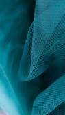 A close-up of teal fabric featuring a mesh-like texture with small, repetitive holes, creating a delicate and airy appearance.