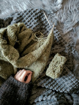 A hand holds a knitting project made of green yarn, lying on various textured blankets. Nearby rests a skein of yarn and knitting needles.