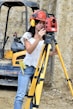 A woman wearing a red safety helmet and yellow earmuffs operates a surveying instrument mounted on a tripod. She is standing in a construction area with a compact excavator in the background. Her casual attire includes a white eyelet blouse and blue jeans, and she is holding a digital level.