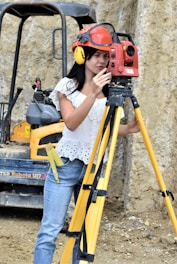 A woman wearing a red safety helmet and yellow earmuffs operates a surveying instrument mounted on a tripod. She is standing in a construction area with a compact excavator in the background. Her casual attire includes a white eyelet blouse and blue jeans, and she is holding a digital level.