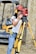 A woman wearing a red safety helmet and yellow earmuffs operates a surveying instrument mounted on a tripod. She is standing in a construction area with a compact excavator in the background. Her casual attire includes a white eyelet blouse and blue jeans, and she is holding a digital level.
