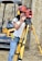 A woman wearing a red safety helmet and yellow earmuffs operates a surveying instrument mounted on a tripod. She is standing in a construction area with a compact excavator in the background. Her casual attire includes a white eyelet blouse and blue jeans, and she is holding a digital level.