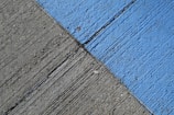 A concrete surface is painted with diagonal lines, featuring a section in blue and another in gray. The texture is rough, with visible grooves and small debris scattered on the surface.