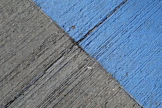 A concrete surface is painted with diagonal lines, featuring a section in blue and another in gray. The texture is rough, with visible grooves and small debris scattered on the surface.