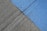 A concrete surface is painted with diagonal lines, featuring a section in blue and another in gray. The texture is rough, with visible grooves and small debris scattered on the surface.