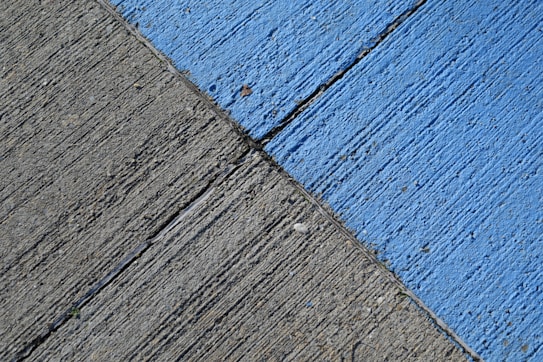 A concrete surface is painted with diagonal lines, featuring a section in blue and another in gray. The texture is rough, with visible grooves and small debris scattered on the surface.