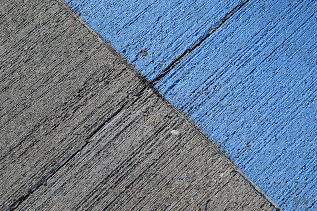 A concrete surface is painted with diagonal lines, featuring a section in blue and another in gray. The texture is rough, with visible grooves and small debris scattered on the surface.