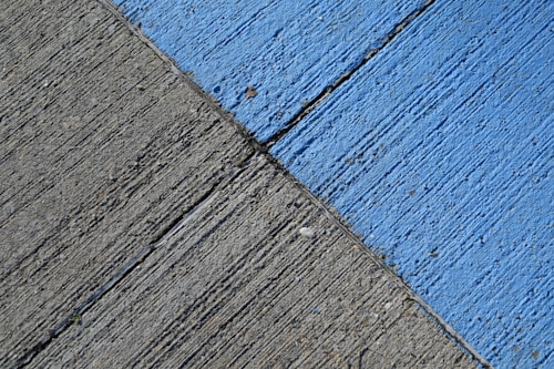 A concrete surface is painted with diagonal lines, featuring a section in blue and another in gray. The texture is rough, with visible grooves and small debris scattered on the surface.