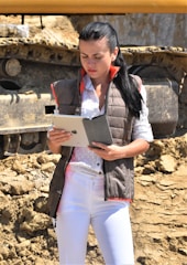 A woman is standing outdoors on construction site soil, wearing a brown vest with orange details, a white polka-dotted shirt, and white pants. She is looking at a tablet and appears focused. Behind her is a large piece of construction machinery with rugged tracks.
