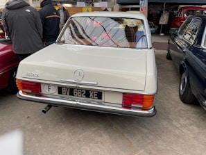 A vintage Mercedes-Benz 200 car with a cream-colored exterior is parked indoors. The car is surrounded by other vehicles and several people, some of whom are examining the cars. The license plate is visible, and there are stickers on the rear indicating it is from 1975. The setting seems to be an indoor car exhibition or show.