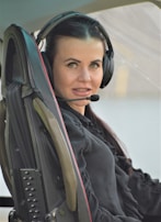 A woman with long dark hair wearing a headset with a microphone is seated inside a vehicle. She wears a black blouse and is looking directly at the camera.