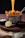 Golden waffle fries are served in a metal basket with melted cheese being poured over them. The basket is placed on a wooden surface, accompanied by a small black bowl of white creamy dip, a black bowl of ketchup, and a half lemon.