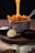 Golden waffle fries are served in a metal basket with melted cheese being poured over them. The basket is placed on a wooden surface, accompanied by a small black bowl of white creamy dip, a black bowl of ketchup, and a half lemon.