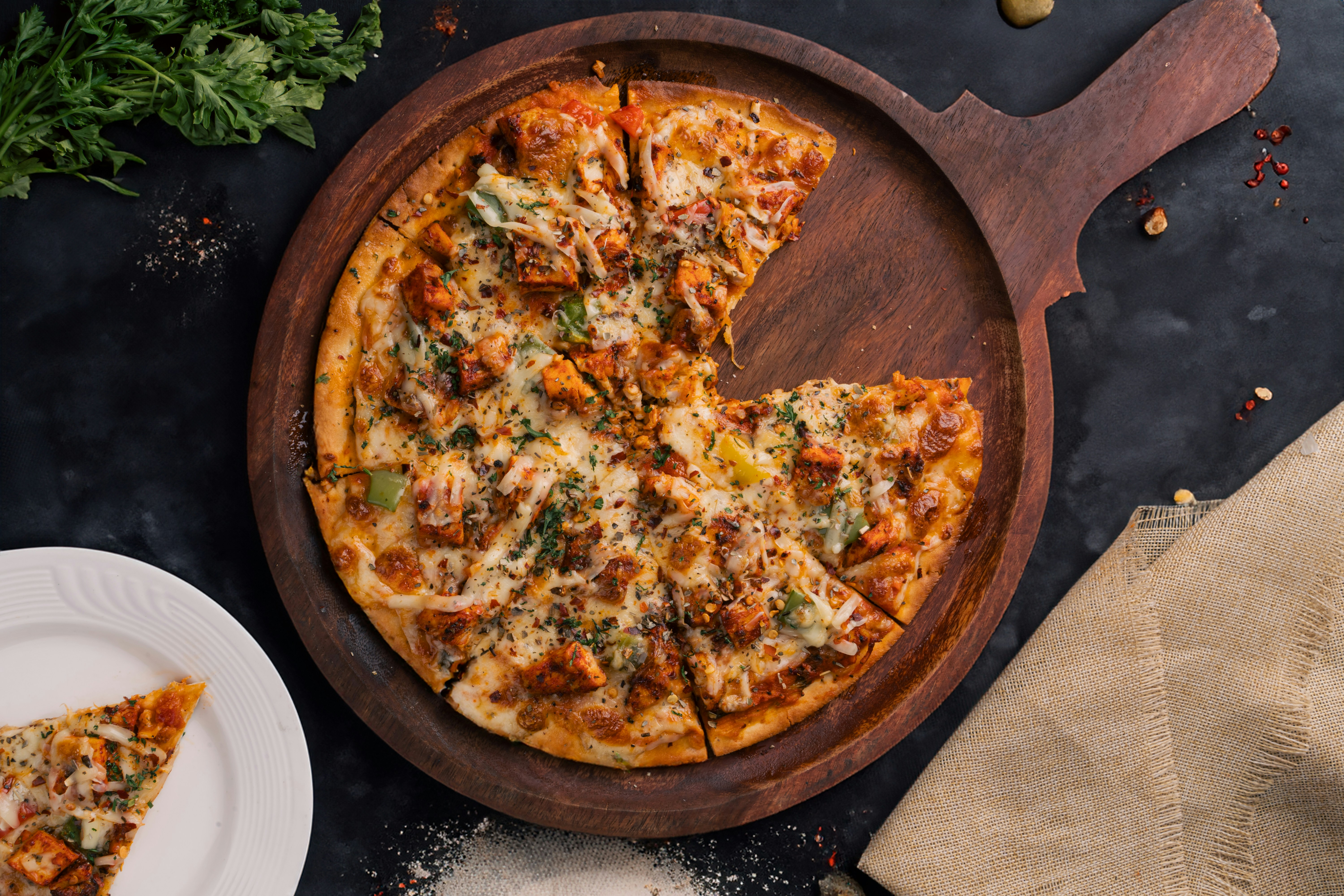 Freshly baked wood-fired chicken pizza topped with melted cheese, herbs, and vegetables on a rustic wooden board with a slice served on a white plate.