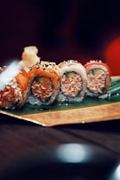 A set of sushi rolls placed on a leaf atop a decorative rectangular tray. The rolls feature a variety of colorful toppings including raw fish, sesame seeds, and avocado. A blurred background adds depth to the image, emphasizing the sushi as the focal point.