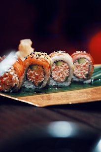 A set of sushi rolls placed on a leaf atop a decorative rectangular tray. The rolls feature a variety of colorful toppings including raw fish, sesame seeds, and avocado. A blurred background adds depth to the image, emphasizing the sushi as the focal point.