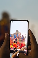 A person is holding a smartphone, capturing an image of a colorful statue of a deity. The deity has multiple arms and is adorned with vibrant decorations and garlands. The scene is lively, suggesting a festival or celebration, with a crowd gathered around the statue.