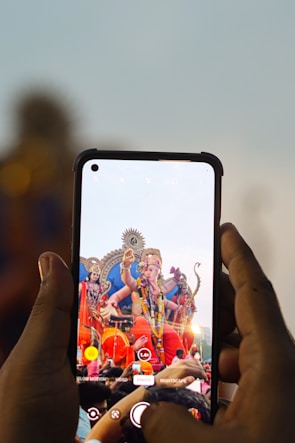A person is holding a smartphone, capturing an image of a colorful statue of a deity. The deity has multiple arms and is adorned with vibrant decorations and garlands. The scene is lively, suggesting a festival or celebration, with a crowd gathered around the statue.