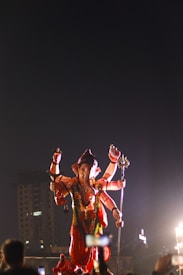 A large statue of the Hindu deity Ganesha, depicted with multiple arms and adorned with traditional attire and garlands, stands prominently against a dark sky. The deity holds various objects symbolizing different attributes. The statue is illuminated by bright lights, with several people capturing the scene with their phones. The background includes a partially visible building.