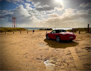 A red convertible sports car is parked on a sandy beach area with tire marks leading towards the water. The sky is partly cloudy, with sunlight breaking through and illuminating the scene. A red metal structure, resembling a lifeguard tower or beacon, and several wooden posts are visible along the edge of the sand dunes. The ocean waves are gently approaching the shore, contributing to the tranquil atmosphere.