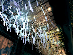 A ceiling installation with numerous white paper strips hanging down, illuminated by lights. The strips have text on them and are part of an artistic display. The arrangement includes delicate fairy lights entwined in a net-like structure.