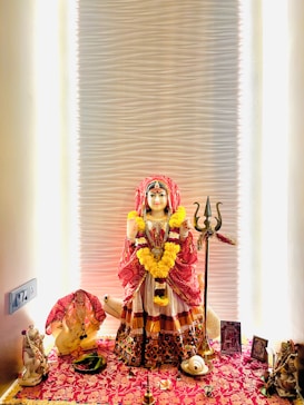 A beautifully decorated shrine featuring a statue dressed in traditional attire with a red and gold theme. The statue is adorned with garlands of yellow flowers and stands on a vibrant floral-patterned cloth. On either side, there are decorative items including small statues, framed images, a trident, and other ritualistic elements. The background has a softly lit, textured wall that enhances the sacred atmosphere.