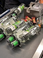 Several clear glass bottles with green labels and black caps are laid out on a conveyor belt along with some small wrapped candy bars. A cardboard box with an image of a bottle is also present.