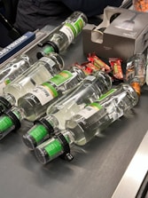 Several clear glass bottles with green labels and black caps are laid out on a conveyor belt along with some small wrapped candy bars. A cardboard box with an image of a bottle is also present.