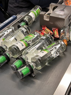 Several clear glass bottles with green labels and black caps are laid out on a conveyor belt along with some small wrapped candy bars. A cardboard box with an image of a bottle is also present.