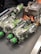 Several clear glass bottles with green labels and black caps are laid out on a conveyor belt along with some small wrapped candy bars. A cardboard box with an image of a bottle is also present.