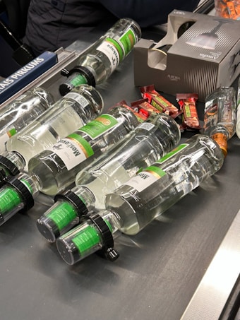 Several clear glass bottles with green labels and black caps are laid out on a conveyor belt along with some small wrapped candy bars. A cardboard box with an image of a bottle is also present.