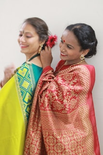Two women dressed in traditional attire are enjoying a moment together. One is wearing a vibrant green and yellow sari, while the other is in a red and gold sari. They are smiling as the woman in the red sari adjusts or pins a decorative red flower in the hair of the woman in green.