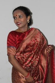 A woman is wearing a traditional red and gold saree with intricate patterns. She has a bindi on her forehead and is smiling gently. Her earrings are large and ornamental, and she wears a gold necklace. Her hair is neatly tied back.