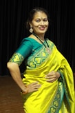 A person is wearing a vibrant yellow and teal traditional saree with intricate patterns. The background consists of dark curtains, suggesting an indoor setting. The person is smiling and appears to be posing elegantly, adorned with jewelry, including earrings and a necklace.