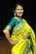 A person is wearing a vibrant yellow and teal traditional saree with intricate patterns. The background consists of dark curtains, suggesting an indoor setting. The person is smiling and appears to be posing elegantly, adorned with jewelry, including earrings and a necklace.