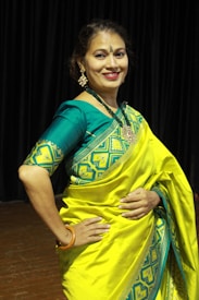 A person is wearing a vibrant yellow and teal traditional saree with intricate patterns. The background consists of dark curtains, suggesting an indoor setting. The person is smiling and appears to be posing elegantly, adorned with jewelry, including earrings and a necklace.