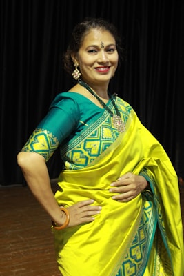 A person is wearing a vibrant yellow and teal traditional saree with intricate patterns. The background consists of dark curtains, suggesting an indoor setting. The person is smiling and appears to be posing elegantly, adorned with jewelry, including earrings and a necklace.