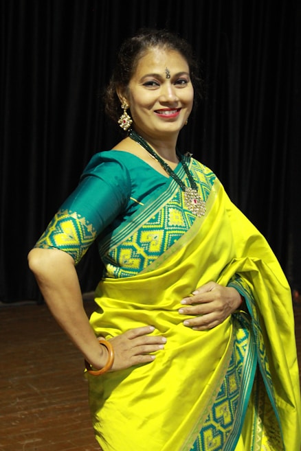A person is wearing a vibrant yellow and teal traditional saree with intricate patterns. The background consists of dark curtains, suggesting an indoor setting. The person is smiling and appears to be posing elegantly, adorned with jewelry, including earrings and a necklace.