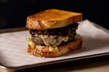 A gourmet sandwich featuring a large, juicy burger patty topped with melted cheese, crispy fried onions, and fresh lettuce, all between two toasted sesame-seed bread slices. The sandwich is placed on a lined tray, set against a dark background.