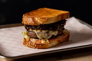 A gourmet sandwich featuring a large, juicy burger patty topped with melted cheese, crispy fried onions, and fresh lettuce, all between two toasted sesame-seed bread slices. The sandwich is placed on a lined tray, set against a dark background.